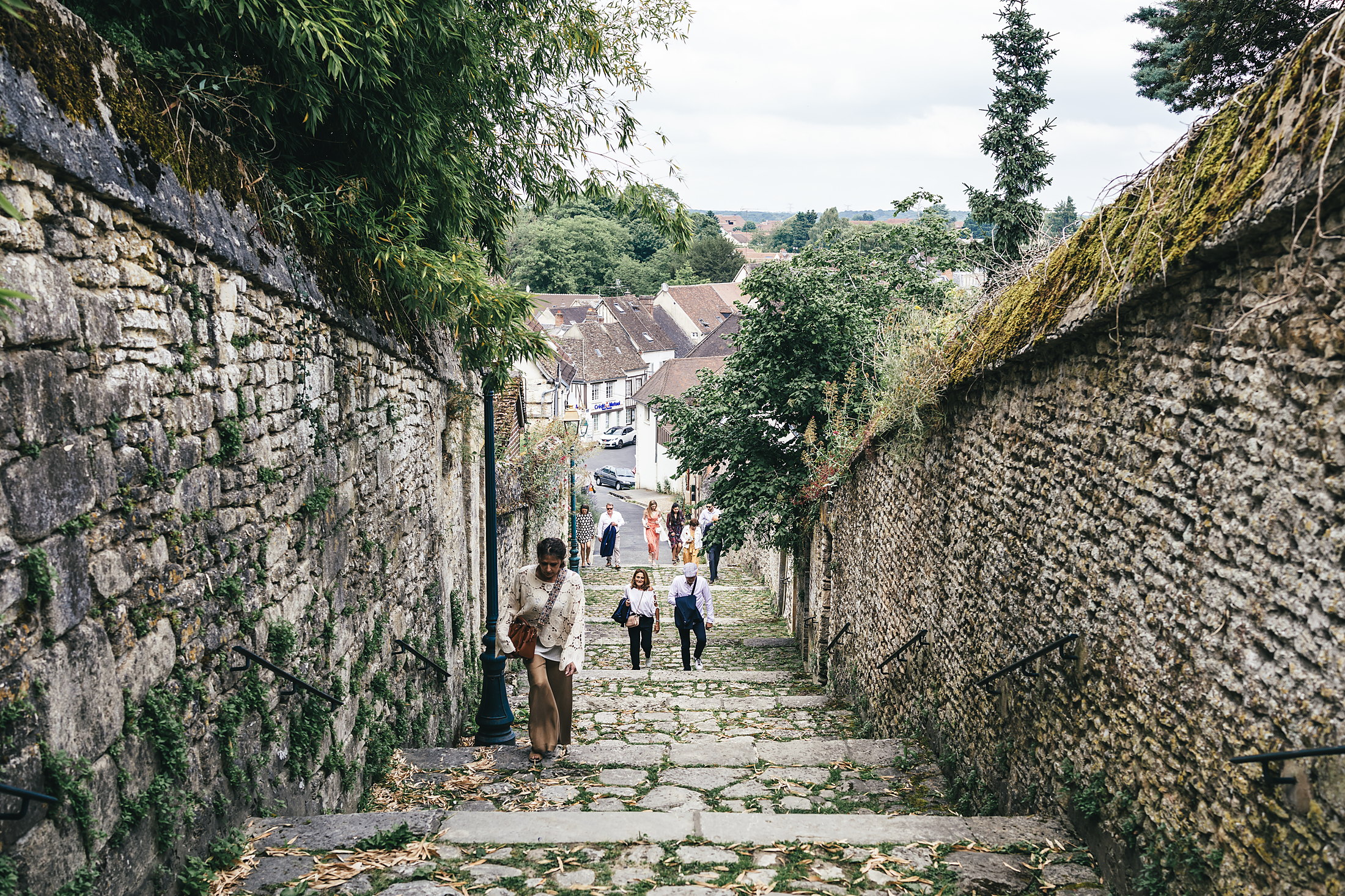 Le cortège descend la ruelle pavée bordée de murs en pierre vers l'église de Chaumont-en-Vexin